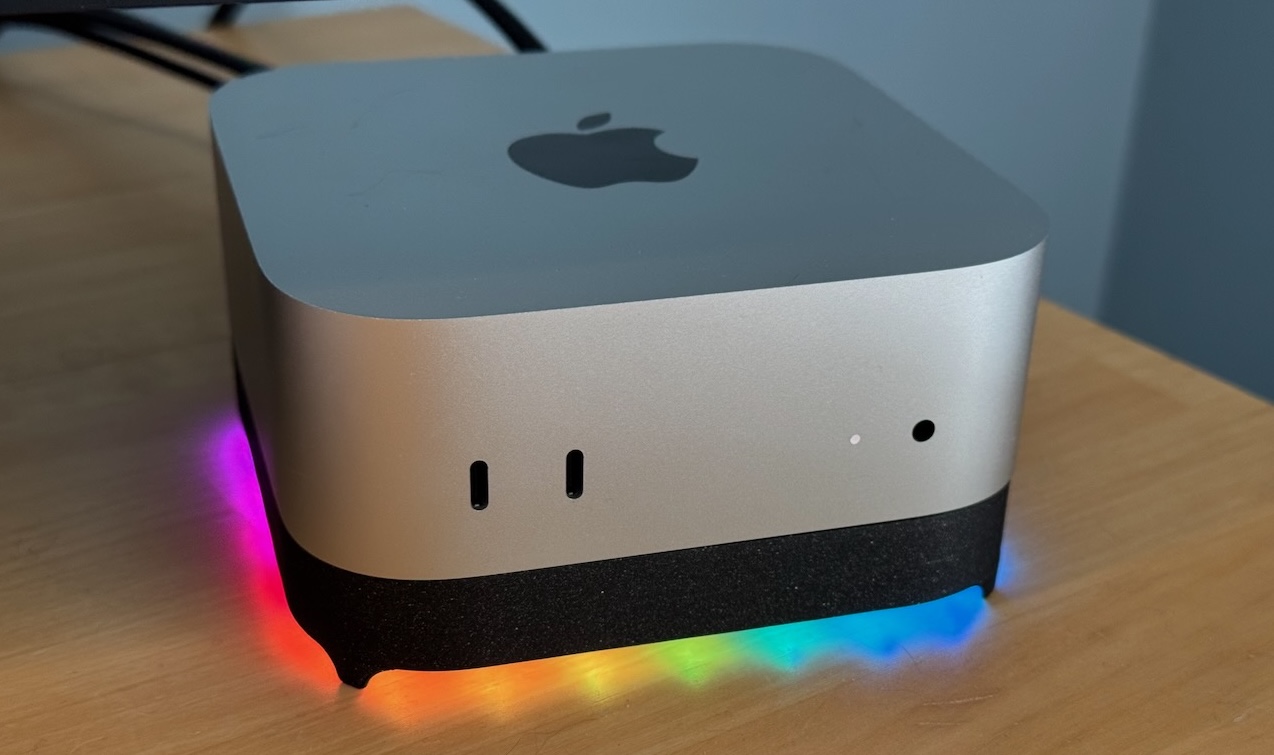 A Mac mini sitting on the black Blinkenhub stand, casting a rainbow of light onto a wooden desk